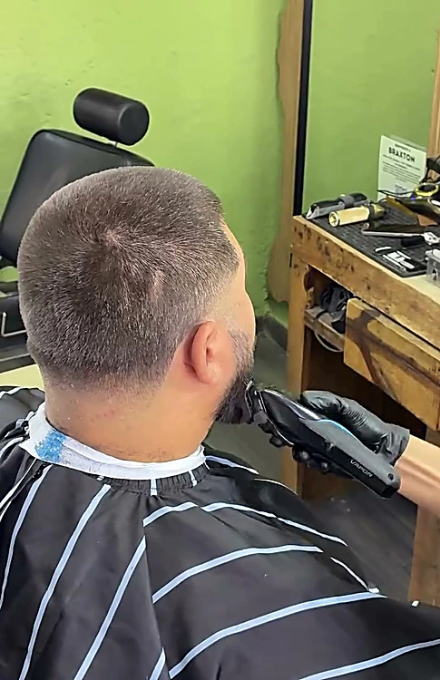 Barbero con guantes negros recorta la barba de un cliente visto de espaldas, muro verde al fondo.