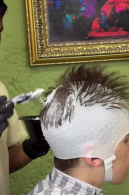 Barbero aplica decoloración con pincel sobre el cabello de un cliente, con gorro de mechas.