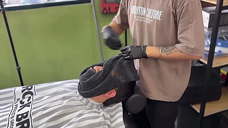 Barbero con playera Braxton Culture cubre la cara de un cliente con una toalla caliente.